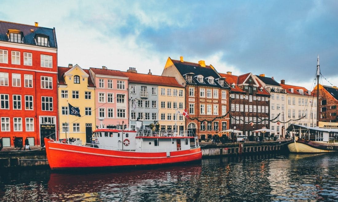 A red hulled boat in a canal with houses behind it.