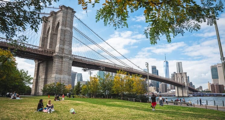 People relaxing on a grassy lawn underneath the Brooklyn Bridge at the Brooklyn Bridge Park in NYC.