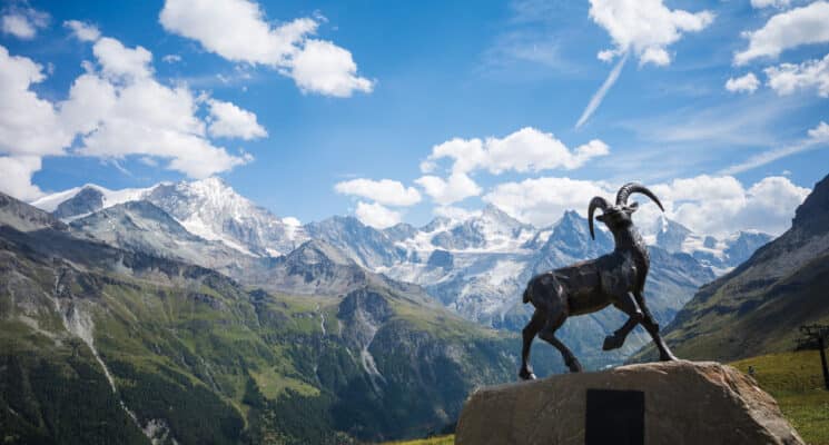 A brass ibex statue stands on a rock, with the majestic Weisshorn and Zinalrothorn mountains in the background.